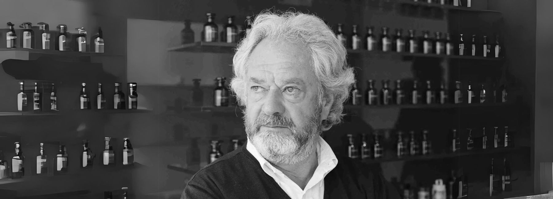 A portrait of perfumer Michel Almairac, shown from the chest up, smiling slightly against a neutral background.